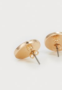 Boucles d'oreilles rondes en métal doré avec une finition texturée, présentant un fermoir à tige et des détails de boucle décoratifs autour du clou.