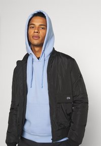 Giacca bomber nera con texture trapuntata, tasche con zip e un felpa blu sotto, dotata di cordini e una tasca frontale.
