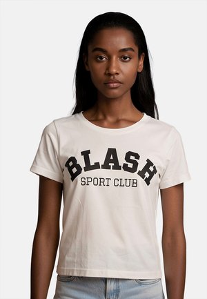Donna con capelli lunghi e scuri indossa una t-shirt bianca con scritta nera "BLASH SPORT CLUB", abbinata a jeans azzurri, rivolta in avanti su sfondo bianco.