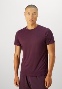 Homme portant un t-shirt de sport moulant bordeaux et un short assorti, se tenant contre un fond clair uni, face à l'avant.