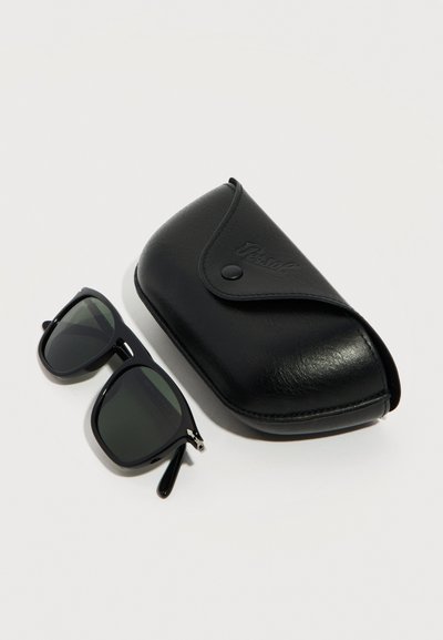 Μαύρα ορθογώνια γυαλιά ηλίου με σκούρους φακούς τοποθετημένα δίπλα σε μια κλειστή μαύρη δερμάτινη θήκη με κουμπί κλιπ.