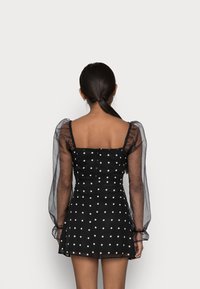 Czarna sukienka w kropki z przezroczystymi puffowanymi rękawami, kwadratowym dekoltem, dopasowanym górą i rozkloszowaną spódnicą. Teksturowany materiał o gładkim wykończeniu.