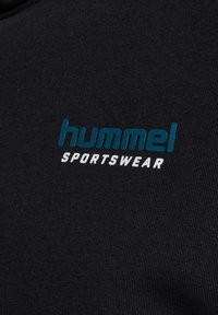 Tissu noir avec le texte « hummel » en bleu et « SPORTSWEAR » en blanc imprimés dessus, montrant le logo de la marque sur le matériau du vêtement.