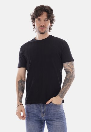 Uomo con capelli ricci che indossa una maglietta nera e jeans blu, mostrando tatuaggi dettagliati su entrambe le braccia su uno sfondo chiaro e uniforme.