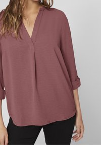 Blouse à manches longues de couleur prune, dotée d'un col en V et d'une coupe décontractée, avec des manches retroussées et une texture subtile. Pantalon noir visible.