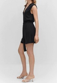 Czarny kombinezon bez rękawów z dekoltem w kształcie V, talią ściągniętą w pasie i luźnymi szortami. Noszony z białymi sandałami na cienkich paskach. Gładka tekstura materiału.