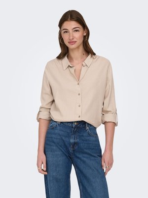 Jeune femme portant une chemise beige à boutons avec manches retroussées et un jean bleu taille haute, debout devant un fond clair uni.