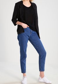 Černý blejzr s uvolněným střihem přes černé tričko, spárovaný s modrými džíny a bílými teniskami. Oblečení má volnočasový design.