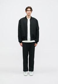 Μαύρο μπουφάν bomber με φερμουάρ, λευκό μπλουζάκι με στρογγυλή λαιμόκοψη και μαύρο παντελόνι. Τα υποδήματα διαθέτουν πολύχρωμες λεπτομέρειες. Απλό φόντο.