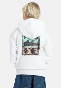 Kind met blond haar in een witte hoodie met een kleurrijk golfontwerp en de tekst "Billabong" op de achterkant, gecombineerd met een blauwe spijkerbroek.