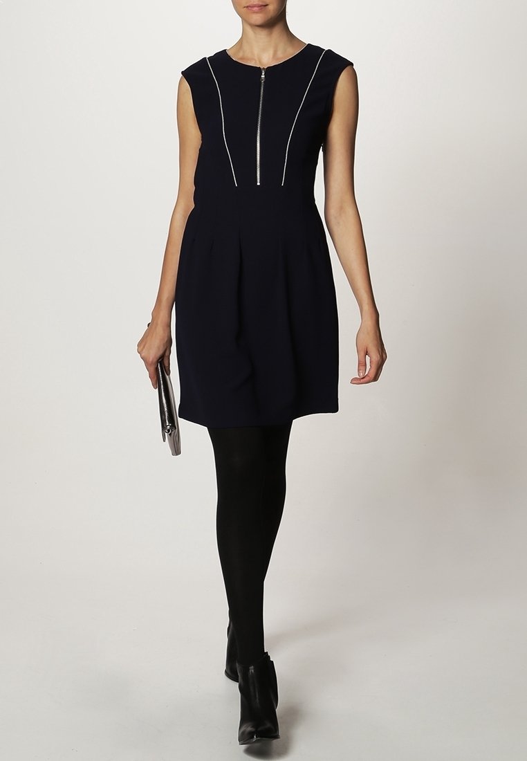 Femme en robe noire sans manches avec un détail de fermeture éclair à l'avant, collants noirs, bottines noires, tenant une petite pochette argentée.