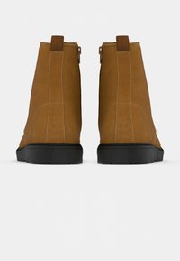 Brązowe zamszowe botki za kostkę o gładkiej fakturze, czarne gumowe podeszwy i małe pętelki z tyłu ułatwiające zakładanie.