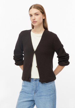 Jeune femme aux cheveux lisses châtain clair porte un cardigan en tricot marron foncé par-dessus une chemise blanche et un jean bleu clair sur fond blanc.