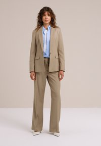 Moteris, stovinti dėvinti smėlio spalvos kostiumą su plačiomis kelnėmis ir šviesiai mėlynę mygtukais užsegamą marškinių palaidinę, dėvi baltus smailius batus, neutralus fonas.