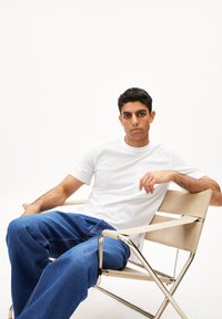 Weißes T-Shirt mit kurzen Ärmeln und Tasche, kombiniert mit locker sitzenden blauen Denim-Jeans, sitzt in einem beigen Stoff-Direktorenstuhl.