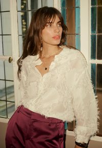 Witte, gestructureerde blouse met rafelige details, gecombineerd met een broek in diep bordeauxrood met hoge taille. Eenvoudige sieraden accentueren de look.