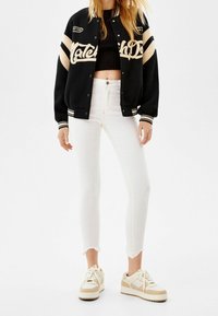 Chaqueta de varsity negra con rayas beige, letras gráficas; combinada con jeans cortos blancos y zapatillas beige con acentos texturizados.