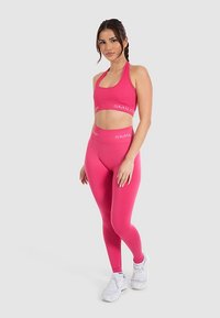 Set composto da reggiseno sportivo rosa e leggings a vita alta con una texture liscia e logo "SMIL2". Indossato con scarpe da ginnastica bianche.