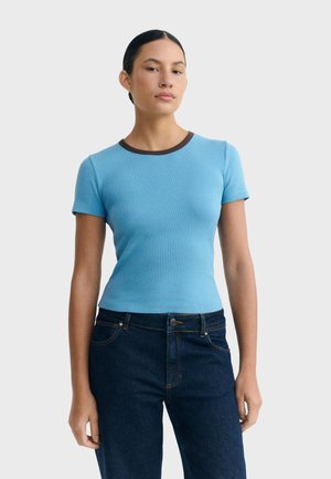 Kobieta o ciemnych włosach związanych do tyłu, ubrana w dopasowany niebieski top z krótkim rękawem i brązowym obszyciem oraz ciemnoniebieskie jeansy, stojąca na tle jednolitego tła.