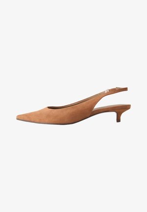 Personne portant un pantalon bleu clair et des chaussures slingback pointues à talons bas beige, debout sur un sol gris.
