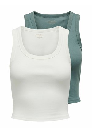 Kobieta z długimi brązowymi włosami, ubrana w biały top na ramiączkach, podarte niebieskie obcisłe dżinsy i czarno-białe trampki, stojąca na jednolitym tle.