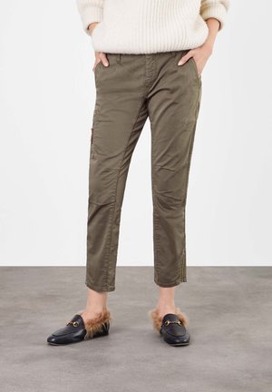 Personne portant un pantalon cargo vert olive avec poches latérales, un pull crème et des mocassins noirs doublés de fourrure, debout sur un sol gris.