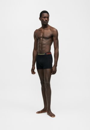 Homme athlétique à la peau foncée debout pieds nus, portant un boxer noir avec "ALLEMAGNE" et "BJÖRN" sur la ceinture, regardant sur le côté.