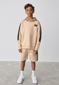 Beige hættetrøje med brun detalje og logo, kombineret med beige shorts med sidelommer og en brun stribe. Hvid sneakers fuldender looket.