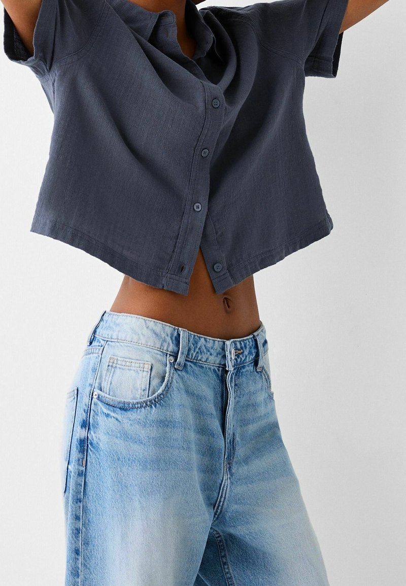 Szara krótko rękawowa bluzka z guzikiem, krótka, zestawiona z jasnobłękitnymi spodniami o wysokim stanie, które mają wybledzoną teksturę i klasyczne kieszenie.