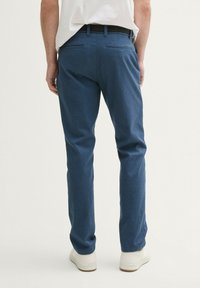 Rückansicht einer Person, die blaue Slim-Fit-Hosen mit aufgesetzten Gesäßtaschen, weiße Sneakers und ein hineingestecktes weißes Hemd trägt.