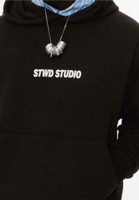 Personne portant un sweat à capuche noir avec le texte "STWD STUDIO" et un collier argenté comportant plusieurs anneaux détaillés.
