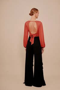 Vrouw met haar in een knot, gekleed in een roestkleurige top met open rug en strik in de taille, en zwarte wijde broek, staand tegen een neutrale achtergrond.