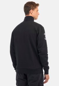 Fekete pulóver magas gallérral, textúrált felülettel és nagy szürke számmal a bal ujján. Puha, laza anyag.