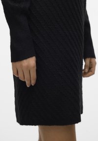 Abito nero lavorato a maglia con un motivo a coste diagonali testurizzato. Maniche lunghe con polsini a coste. L'orlo inferiore è leggermente irregolare.