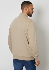 Sweat-shirt beige à demi-zip avec une texture douce et des poignets côtelés. Associé à un jean en denim bleu, soulignant une coupe décontractée.