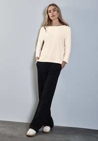 Maglietta a maniche lunghe crema con scollatura a barca, abbinata a pantaloni neri larghi e scarpe da ginnastica bianche con dettagli beige.