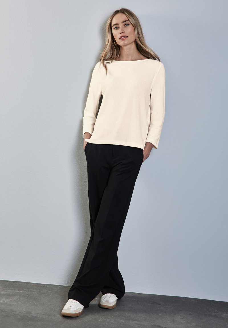 Maglietta a maniche lunghe crema con scollatura a barca, abbinata a pantaloni neri larghi e scarpe da ginnastica bianche con dettagli beige.