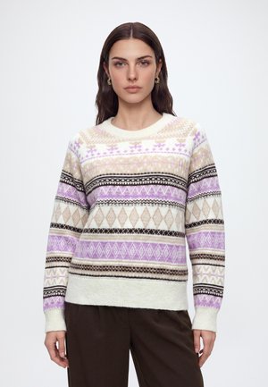 Femme aux longs cheveux bruns portant un pull en maille beige, violet et blanc à motifs et un pantalon marron foncé, debout devant un fond clair uni.