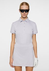 Ljust lila pikétröja med korta ärmar och knappslå, parat med en matchande kjol. Svarta solglasögon och en vit handske accentuerar outfiten.