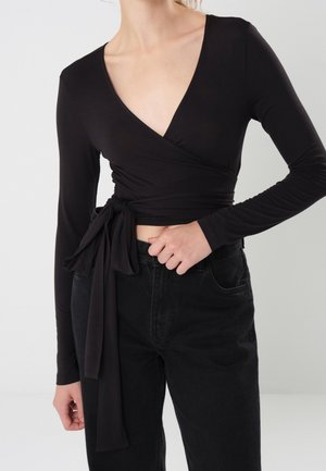 Woman wearing a black long-sleeve wrap crop top tied at the waist paired with high-waisted black jeans against a plain background.