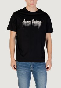 Maglietta nera in cotone a maniche corte con una grafica bianca "Armani Exchange" e un design leggermente testurizzato. Indossata con jeans blu.