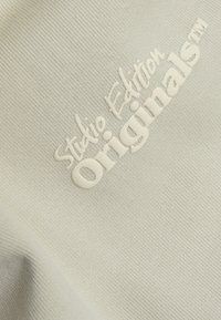 Helles beige Textil mit geprägter Schrift: "Studio Edition Originals™" und einer strukturierten, gerippten Oberfläche.