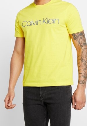 Mężczyzna ubrany w żółtą koszulkę z krótkim rękawem Calvin Klein i czarne dżinsy, z widocznym tatuażem na prawym przedramieniu.