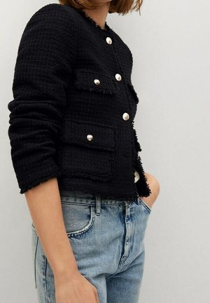 Blazer noir texturé à boutons dorés et poches à rabat, porté avec un jean taille haute bleu clair sur un fond uni.