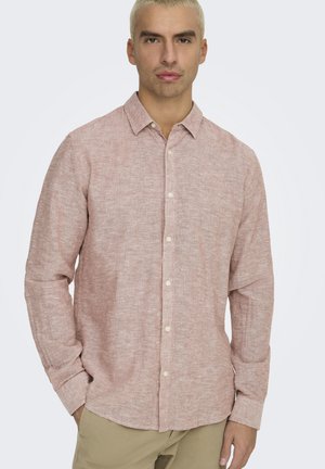 Giovane uomo con capelli biondi corti che indossa una camicia rosa chiaro a maniche lunghe con bottoni e pantaloni beige, in piedi con le mani parzialmente nelle tasche.
