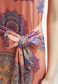 Gros plan d'une ceinture en tissu nouée sur une robe pêche avec des motifs mandala colorés et complexes, montrant une partie d'un bras nu.