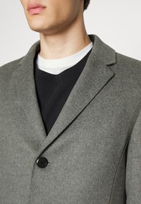 Cappotto di lana grigia con rever a contrasto e un singolo bottone nero, indossato su una maglietta nera a collo rotondo. Texture morbida e vestibilità su misura.