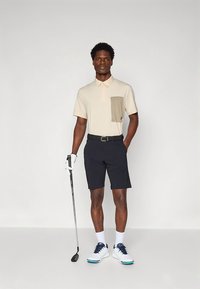 Polo beige con bolsillo marrón claro, combinado con pantalones cortos oscuros; el modelo sostiene un palo de golf; zapatos blancos con detalles azules.
