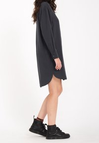 Robe-shirt grise foncée avec des manches longues, fentes sur les côtés, texture douce et ourlet légèrement arrondi. Associée à des bottines noires à lacets.