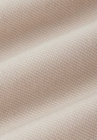 Zbliżenie na beżowy materiał z małym teksturowanym, tkanym wzorem oraz miękkimi zagięciami tworzącymi cienie.